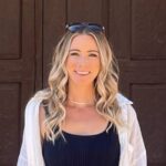 A woman smiling with blonde hair and sunglasses on her head before a dark background